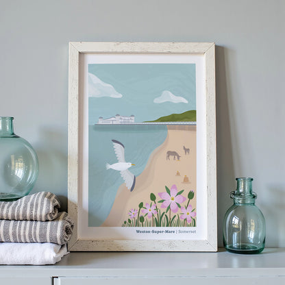 A framed art print featuring a coastal scene labeled 'Western-Super-Mare | Somerset' is displayed on a shelf. The print shows a seagull, donkeys, beach, flowers, and the sea, surrounded by folded towels and glass bottles.