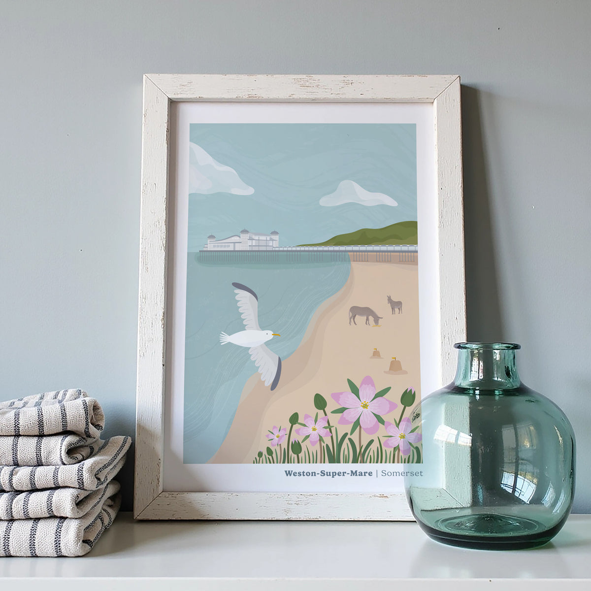 A framed art print depicting the coastal area of Weston Super Mare with a seagull, beach, flowers, and a pier, styled on a shelf next to a folded striped towel and a translucent blue glass bottle against a light grey wall.