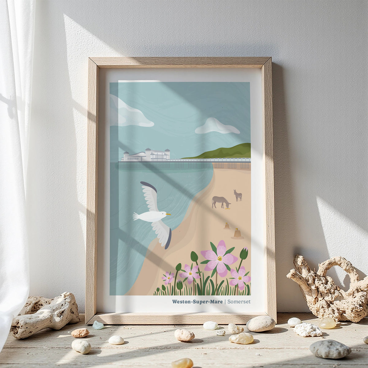A framed art print of Weston-Super-Mare, Somerset, featuring a sea front beach scene with a seagull, donkeys, flowers, and a pier, displayed on a wooden surface with decorative driftwood and pebbles against a white wall.