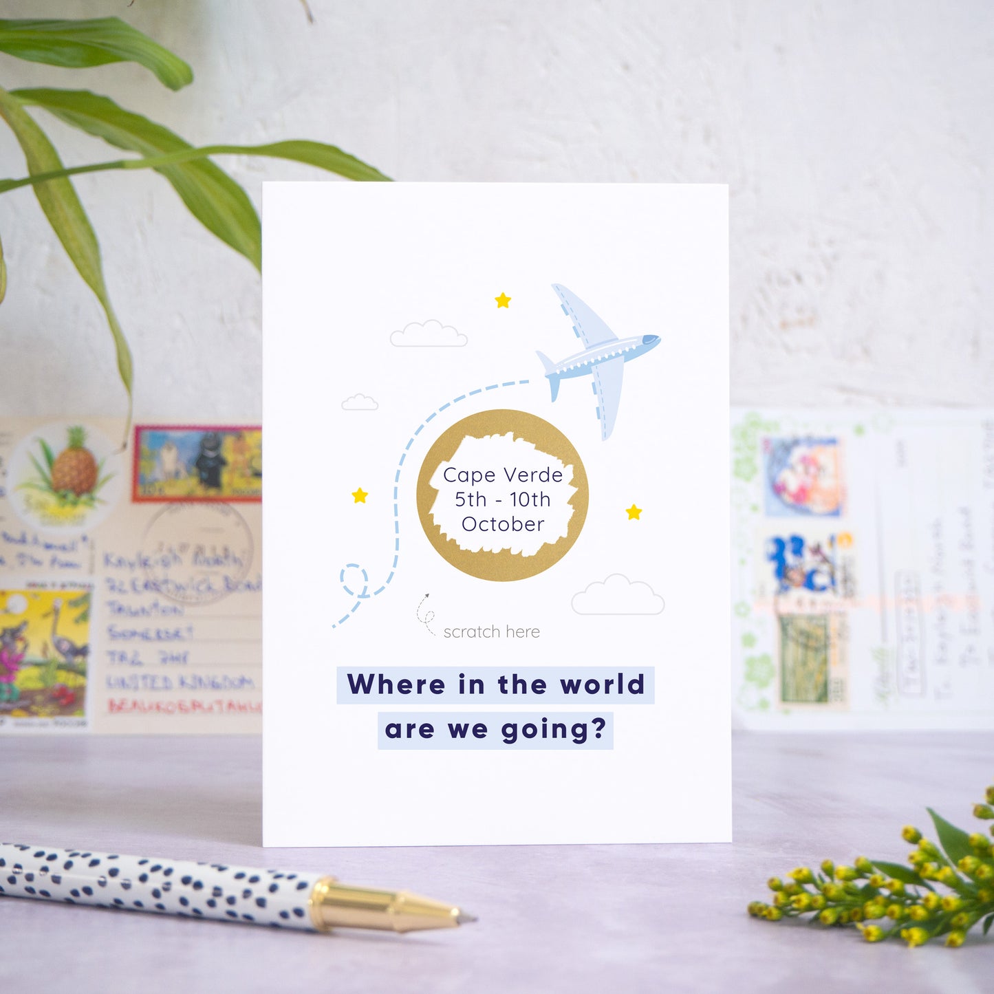 A holiday reveal scratch card featuring a plane flying over the gold scratch panel. The gold panel has been scratched off to reveal the holiday destination. Beneath the plane and scratch panel the text reads: “where in the world are we going”. The card is standing on a grey surface with postcards in the background and a pen and foliage in the foreground. This is the blue colour palette.