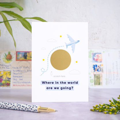 A holiday reveal scratch card featuring a plane flying over the gold scratch panel. The gold panel has not yet been scratched off to reveal the holiday destination. Beneath the plane and scratch panel the text reads: “where in the world are we going”. The card is standing on a grey surface with postcards in the background and a pen and foliage in the foreground.