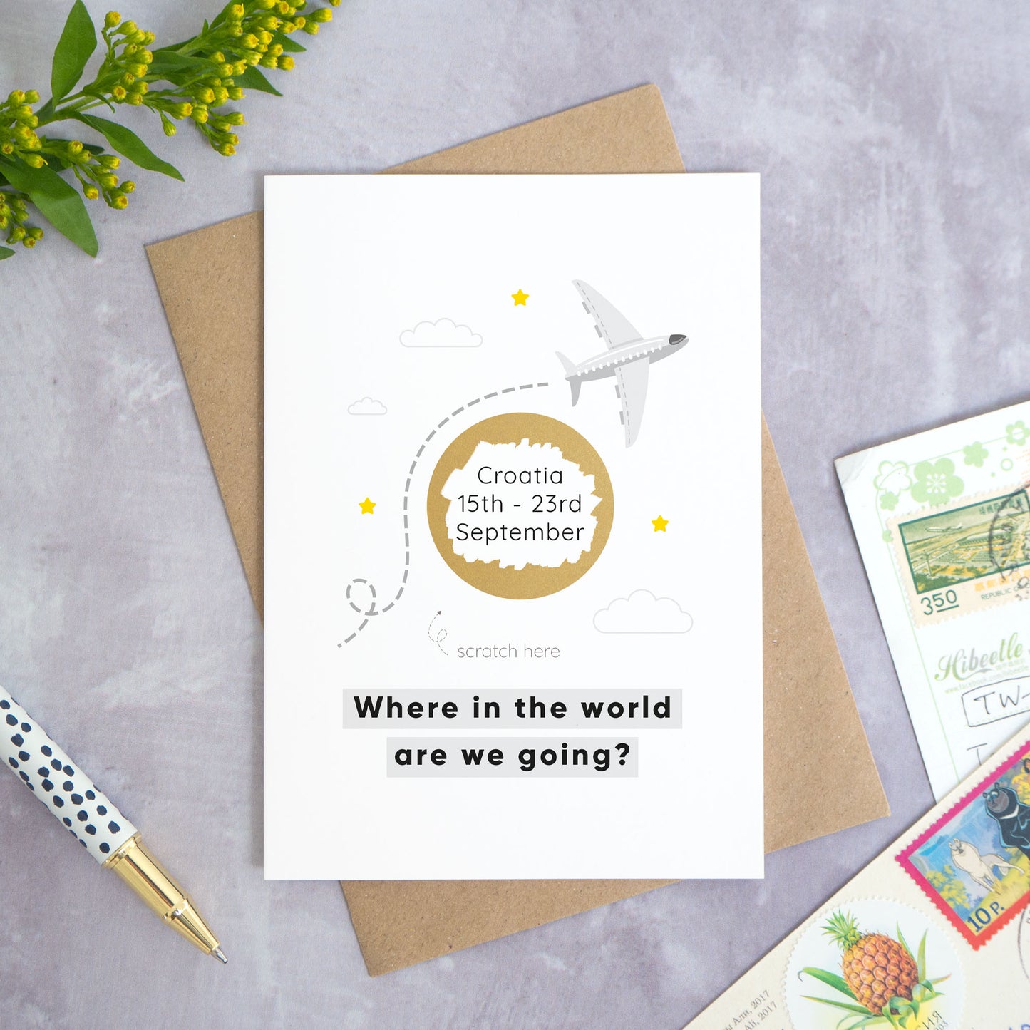 A holiday reveal scratch card featuring a plane flying over the gold scratch panel. The gold panel has been scratched off to reveal the holiday destination. Beneath the plane and scratch panel the text reads: “where in the world are we going”. This card has been shot on a grey background with a pen and foliage. This is the grey colour palette