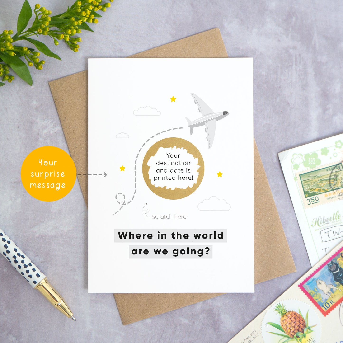 A holiday reveal scratch card featuring a plane flying over the gold scratch panel. The gold panel has been scratched off to reveal the holiday destination. Beneath the plane and scratch panel the text reads: “where in the world are we going”. This card has been shot on a grey background with a pen and foliage. There is also an orange circle pointing to the area which can be personalised. In this case it is the custom message for underneath the scratch panel.