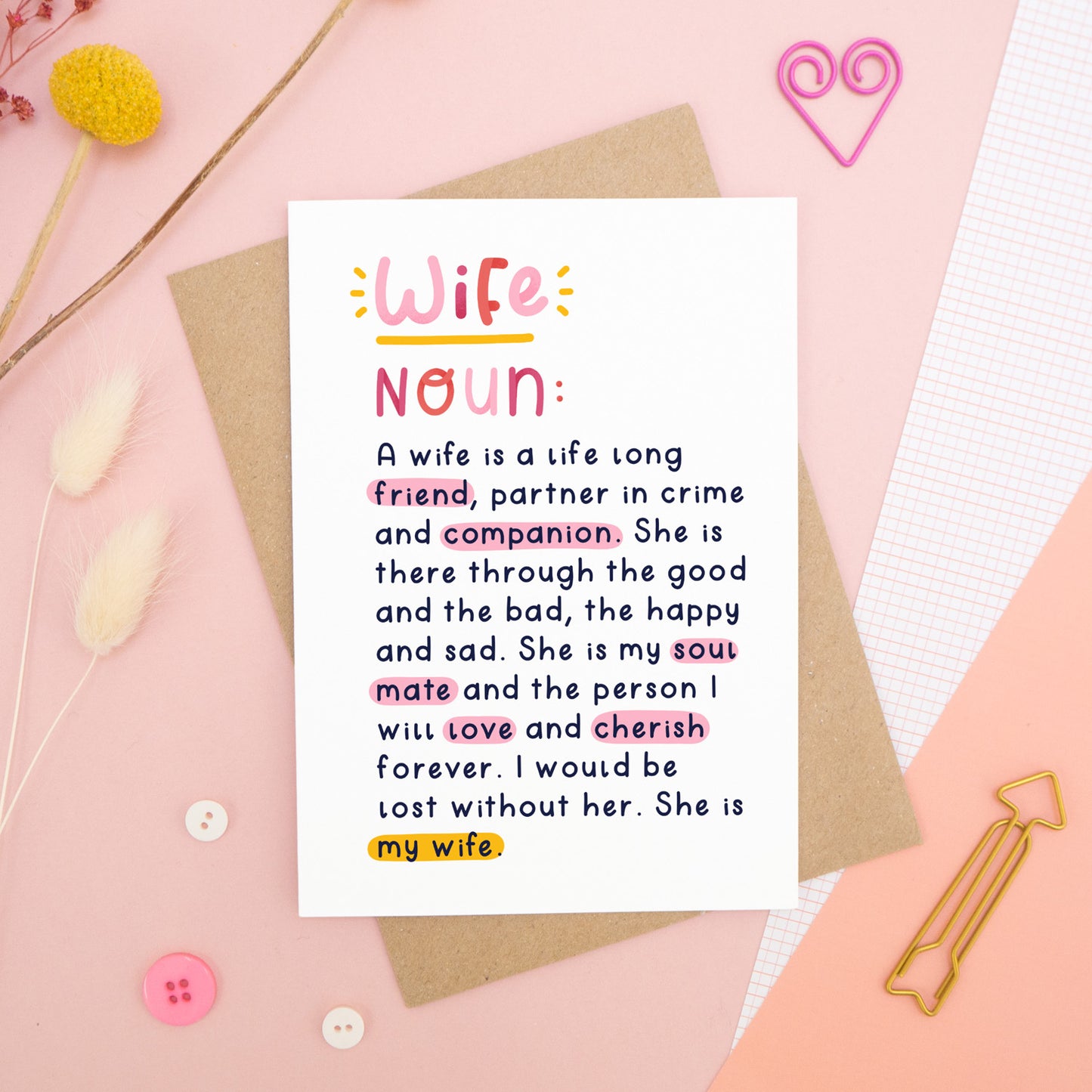 A heartfelt, colourful dictionary definition style card with the definition of a wife, photographed on a pink background with floral props, paper clips, and buttons. 