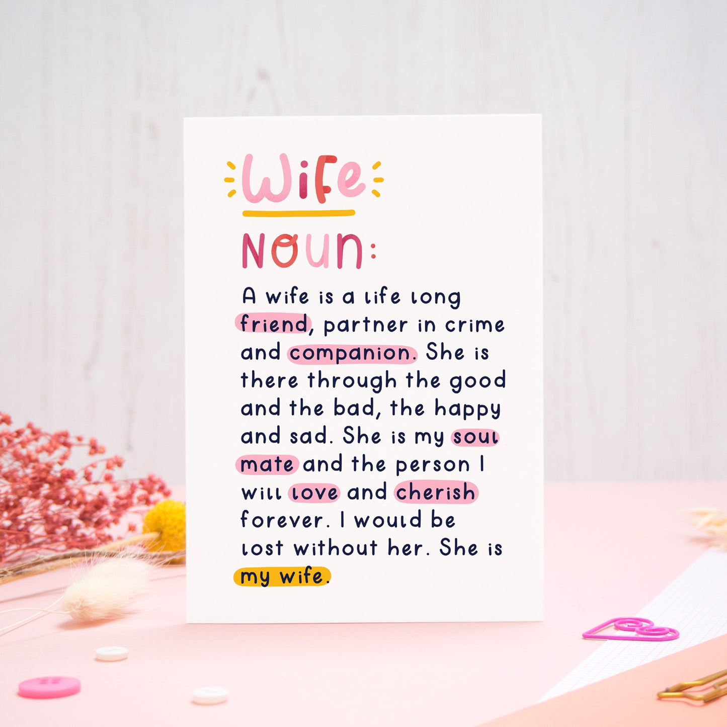 A heartfelt, colourful dictionary definition style card with the definition of a wife, photographed stood on a pink and white background with floral props, paper clips, and buttons. 