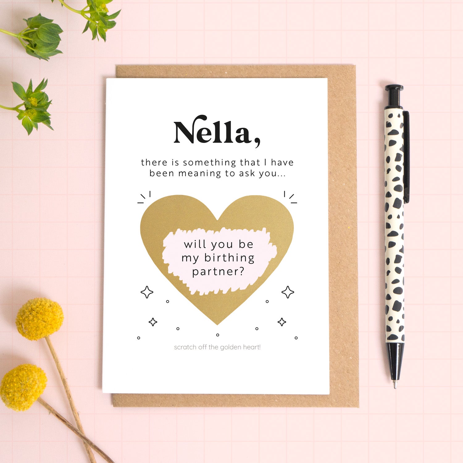 A ‘will you be my birthing partner’ scratch and reveal card photographed lying on a Kraft brown envelope, set upon a pink background surrounded by foliage and a pen for scale. The gold panel has been scratched off to reveal the personalised question. 