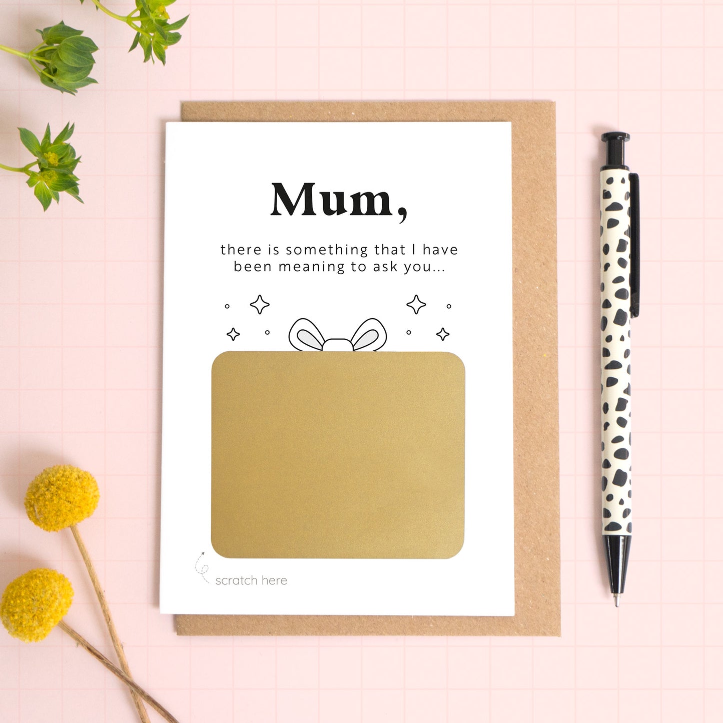 A ‘will you be my birthing partner’  scratch and reveal card photographed lying on a Kraft brown envelope, set upon a pink background surrounded by foliage and a pen for scale. The gold panel has not yet been scratched off. 