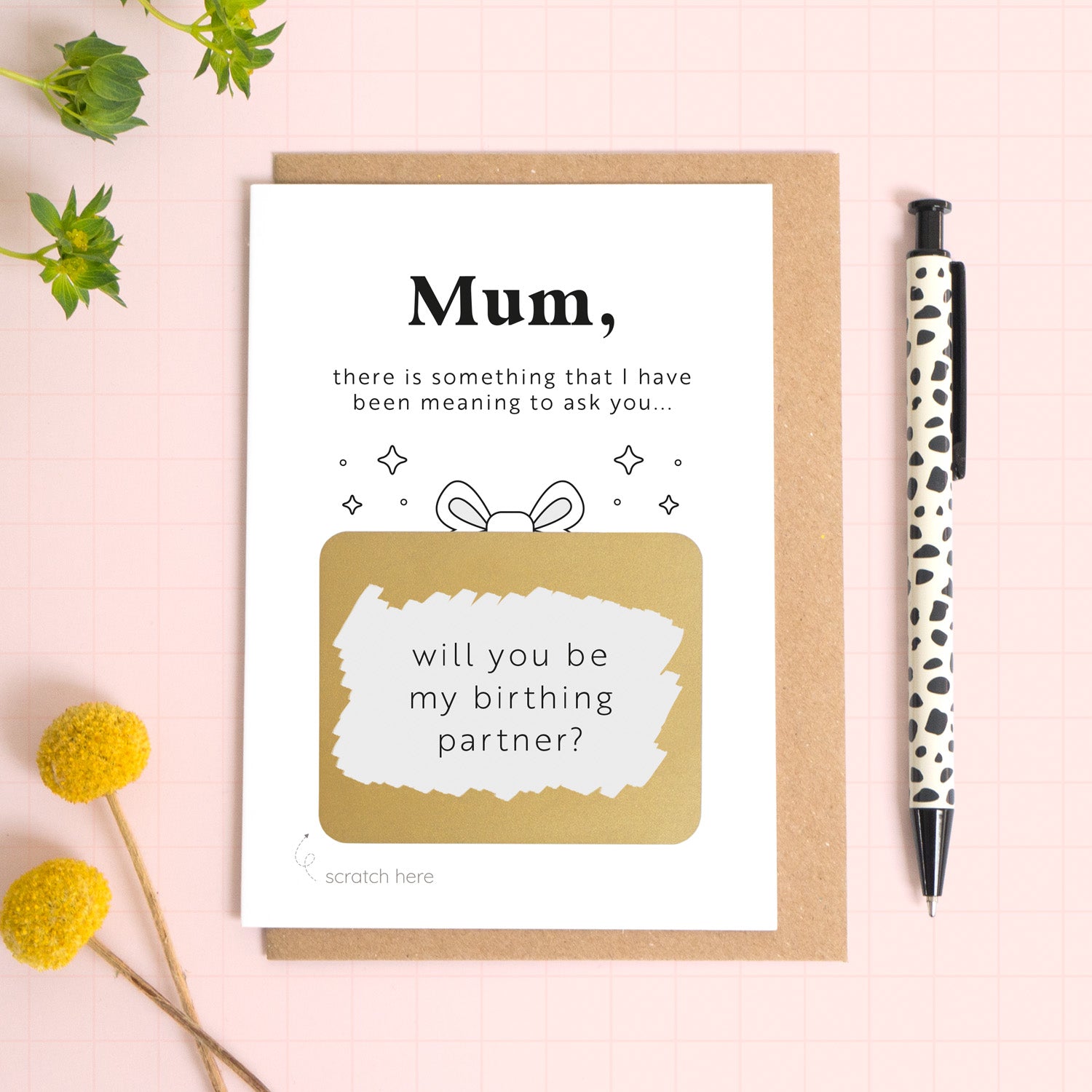 A ‘will you be my birthing partner’ scratch and reveal card photographed lying on a Kraft brown envelope, set upon a pink background surrounded by foliage and a pen for scale. The gold panel has been scratched off to reveal the personalised question. 