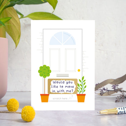 A ‘will you move in with me’ scratch card photographed on a white and grey background with a plant on the left and foliage and a pen on the right. This is the multi-colour version of the card after it has been scratched off.