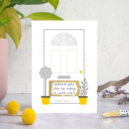 A ‘will you move in with me’ scratch card photographed on a white and grey background with a plant on the left and foliage and a pen on the right. This is the grey version of the card after it has been scratched off.