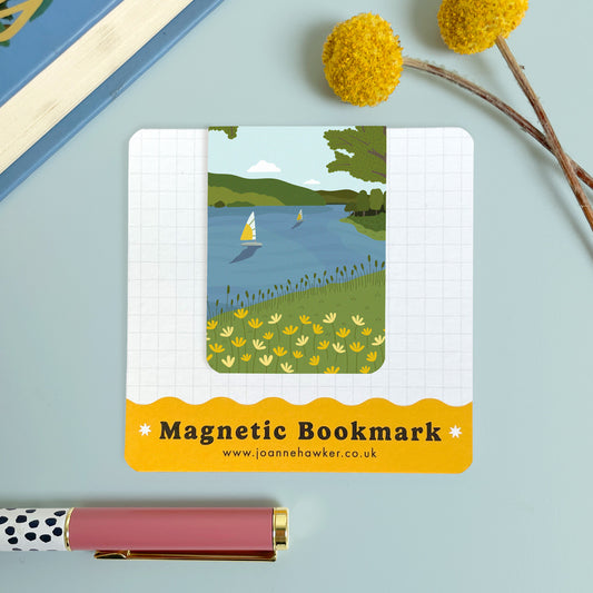 The Wimbleball lake bookmark featuring a view across the Exmoor lake with sailing boats and flowers. The backing card is laid on a light blue surface with a pen and pom pom flowers.