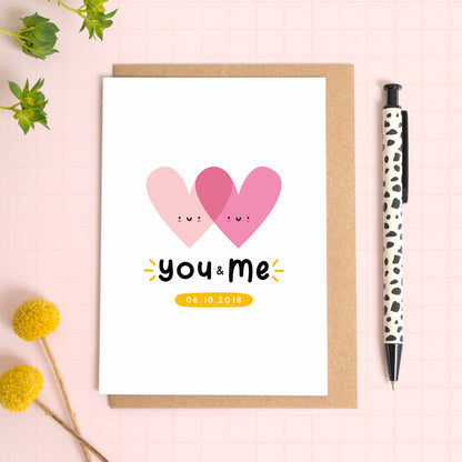 A personalised ‘you and me’ wedding anniversary card with two smiling, overlapping hearts which has been photographed on a Kraft brown envelope upon a pink background surrounded by florals and a pen for scale.