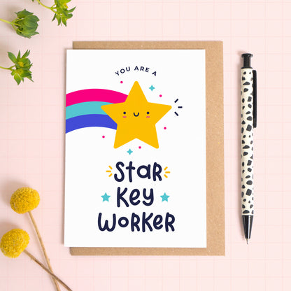 You are a star key worker thank you teacher card with a yellow shooting star that has been photographed on top of a kraft envelope on top of a pink background with foliage and a pen for scale.