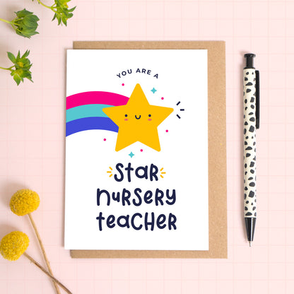 You are a star nursery teacher thank you card with a yellow shooting star that has been photographed on top of a kraft envelope on top of a pink background with foliage and a pen for scale.