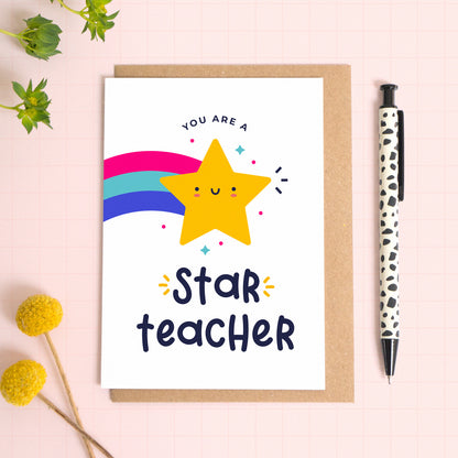 You are a star teacher thank you teacher card with a yellow shooting star that has been photographed on top of a kraft envelope on top of a pink background with foliage and a pen for scale.