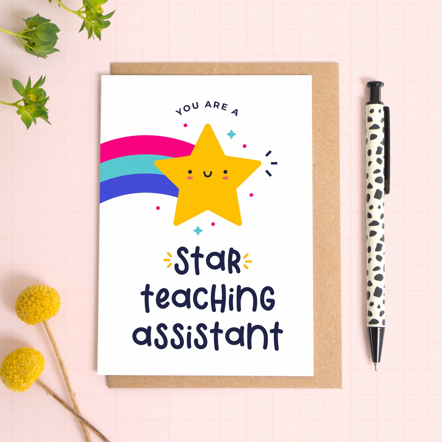 A star teaching assistant thank you teacher card with a yellow shooting star that has been photographed on top of a kraft envelope on top of a pink background with foliage and a pen for scale.