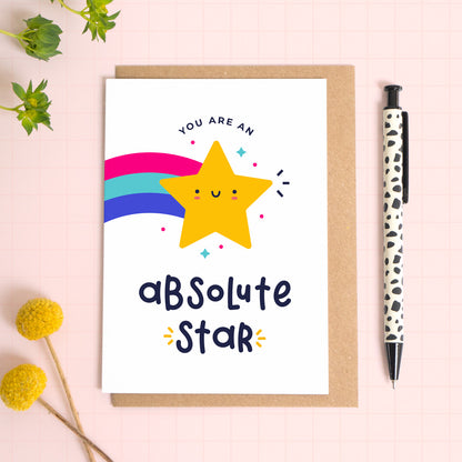 You are an absolute star thank you teacher card with a yellow shooting star that has been photographed on top of a kraft envelope on top of a pink background with foliage and a pen for scale.