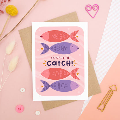 A Valentine’s pun card featuring illustrations of fish and the wording ‘you’re a catch’, photographed on a pink background with floral props, paper clips, and buttons. 