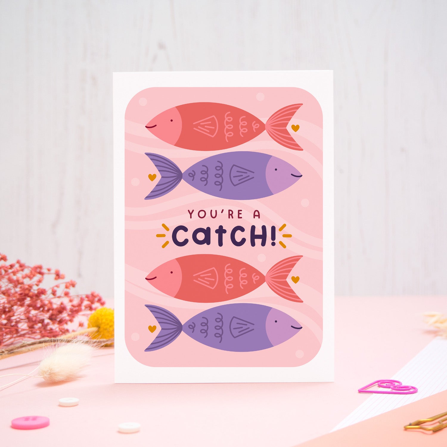 A Valentine’s pun card featuring illustrations of fish and the wording ‘you’re a catch’, photographed stood on a pink and white background with floral props, paper clips, and buttons. 
