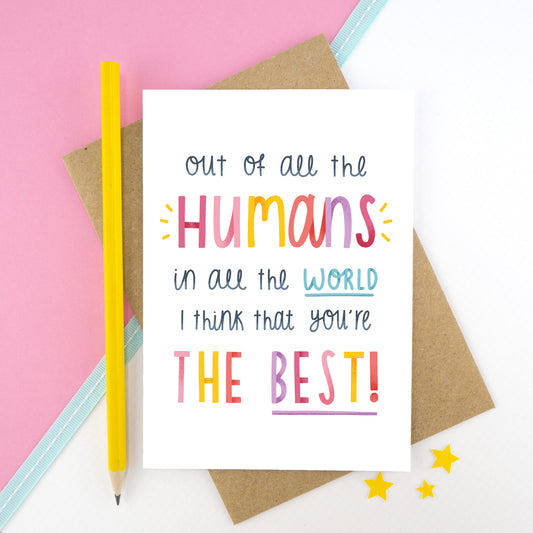 'Out of all the humans in all the world I think that you're the best!' card photographed on a white and pink background with a blue ribbon stripe. There is a yellow pencil to the left and small yellow stars at the bottom.