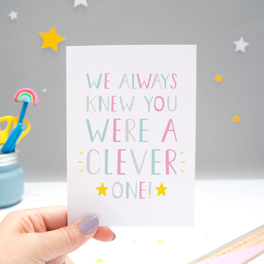 We always knew you were a clever one card held by Joanne Hawker over a grey background with white and yellow stars. The typography is in shades of pink, grey and blue with two yellow stars.