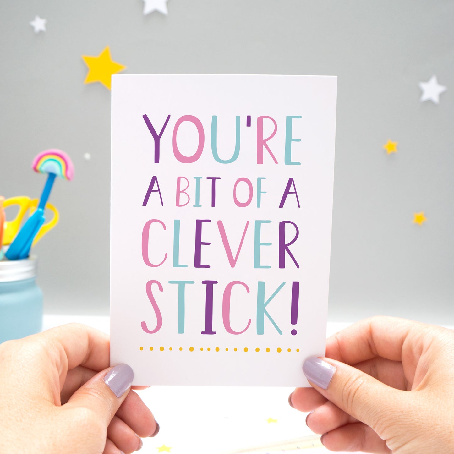 You're a bit of a clever stick card designed and made by Joanne Hawker in her Somerset Studio is being held against a grey background with yellow and white stars. The typography is in varying tones of blue, pink, purple and a pop of yellow!