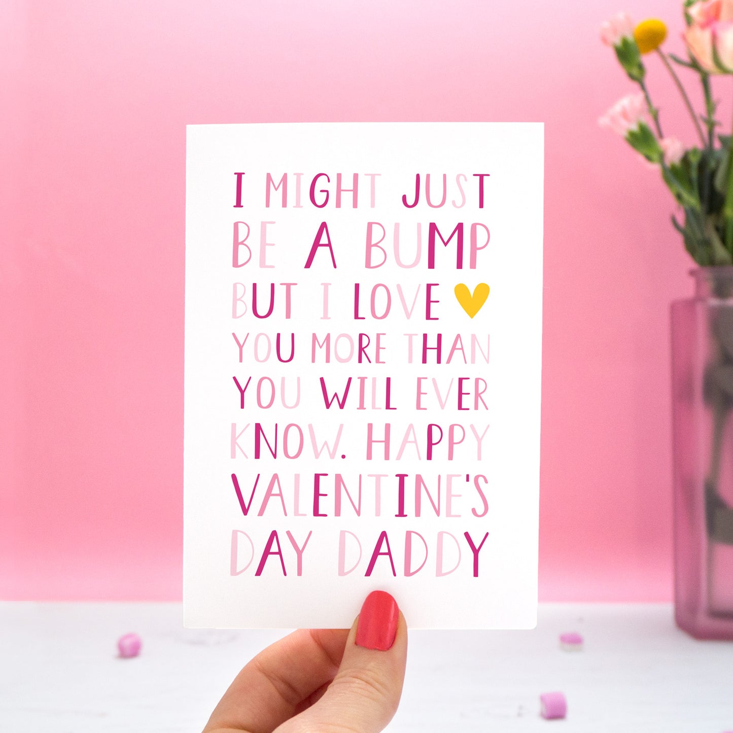 A valentine's card from the 'bump' with text in different shades of pink that reads "I might just be a bump but I love you more thank you will ever know. Happy Valentine's day Daddy." The card is held in one hand and there is a vase of flowers in the pink background.