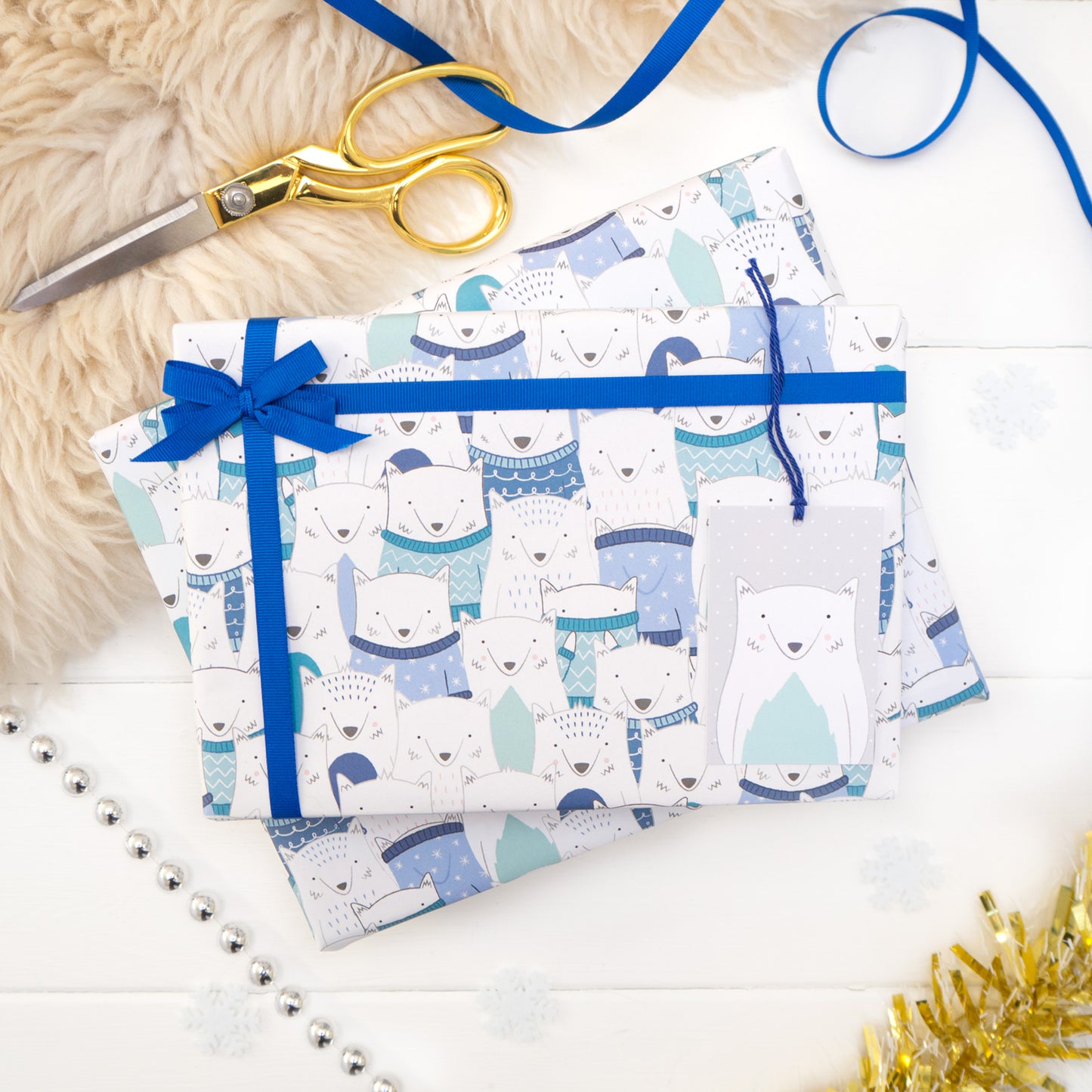 A parade of arctic foxes wearing a variety of woolly jumpers printed onto white gift wrap. Matching fox tag shown on the bottom right of the wrapping paper. The present is strung with blue ribbon and features a small bow.