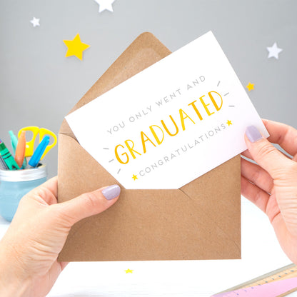 You only went and graduated congratulations card being pulled from an envelope by Joanne Hawker in front of a grey background with white and yellow stars. The typography is a mix of grey and varying tones of yellow and orange.