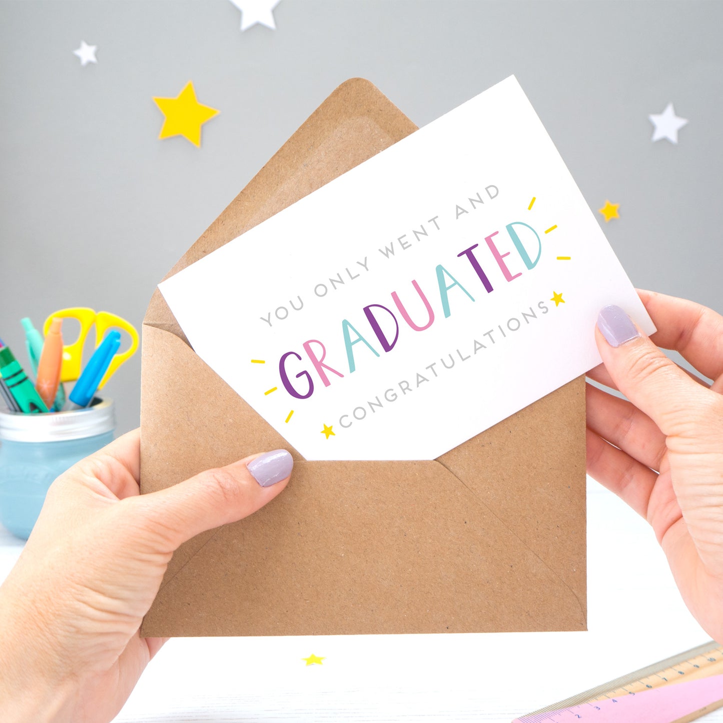 You only went and graduated congratulations card being pulled from an envelope by Joanne Hawker in front of a grey background with white and yellow stars. The typography is a mix of grey and varying tones of blue, pink and purple.