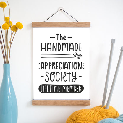 The handmade appreciation society print in black and white and held in a magnetic frame next to a vase of yellow flowers and wool with knitting needles.