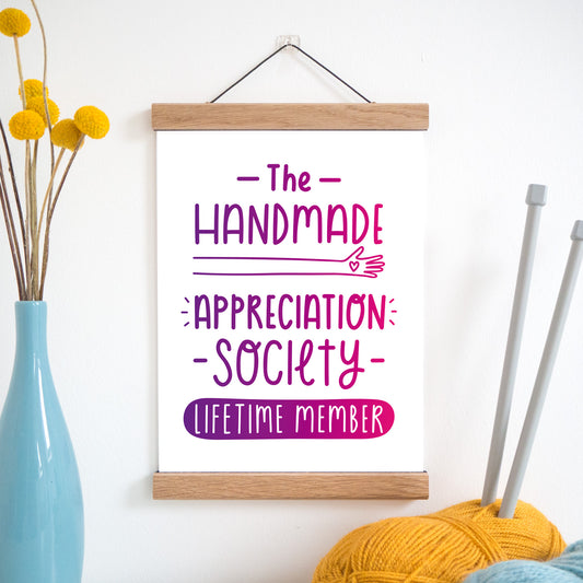 The handmade appreciation society print in purple and pink ombre and held in a magnetic frame next to a vase of yellow flowers and wool with knitting needles.