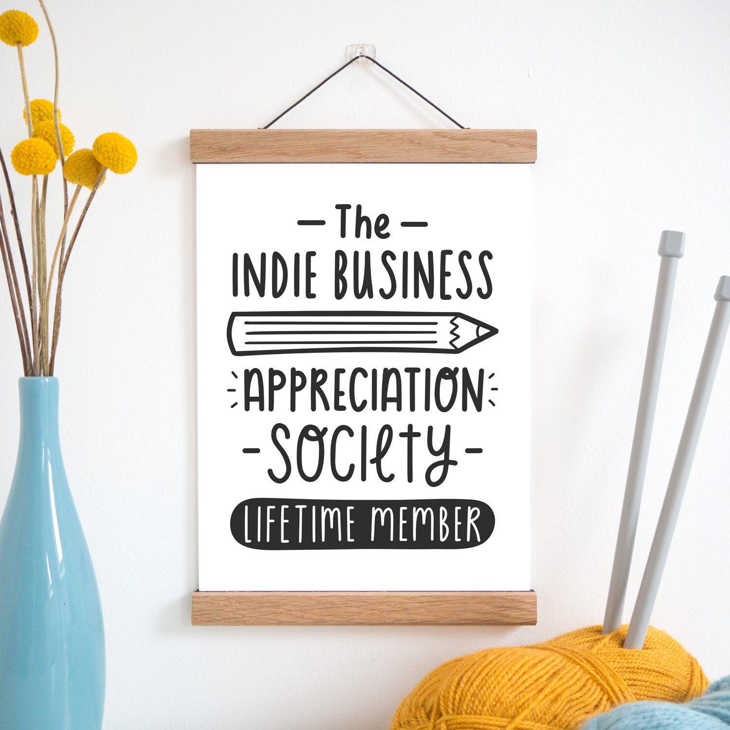 The indie business appreciation society print in black and white and held in a magnetic frame next to a vase of yellow flowers and wool with knitting needles.