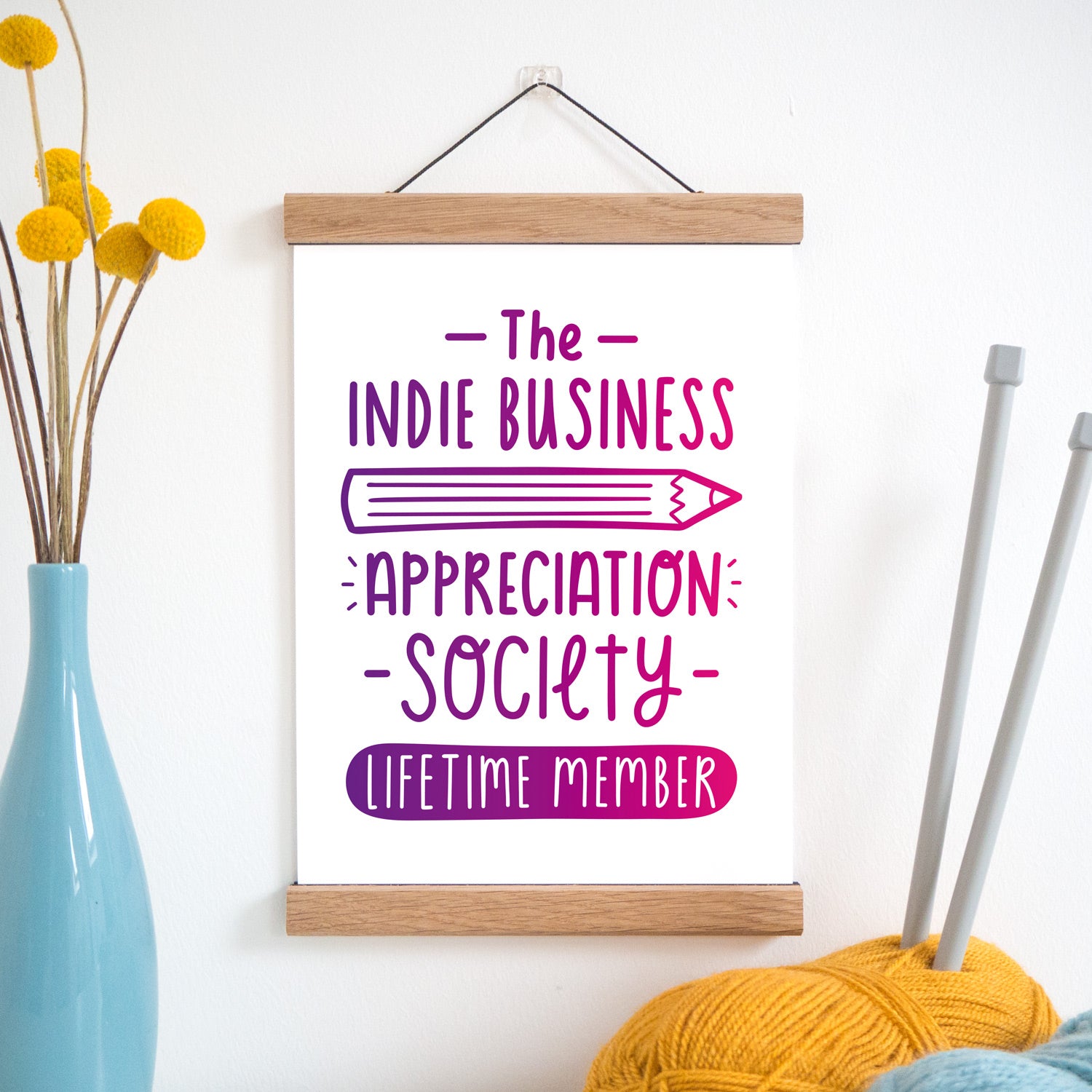 The indie business appreciation society print in pink and purple ombre and held in a magnetic frame next to a vase of yellow flowers and wool with knitting needles.