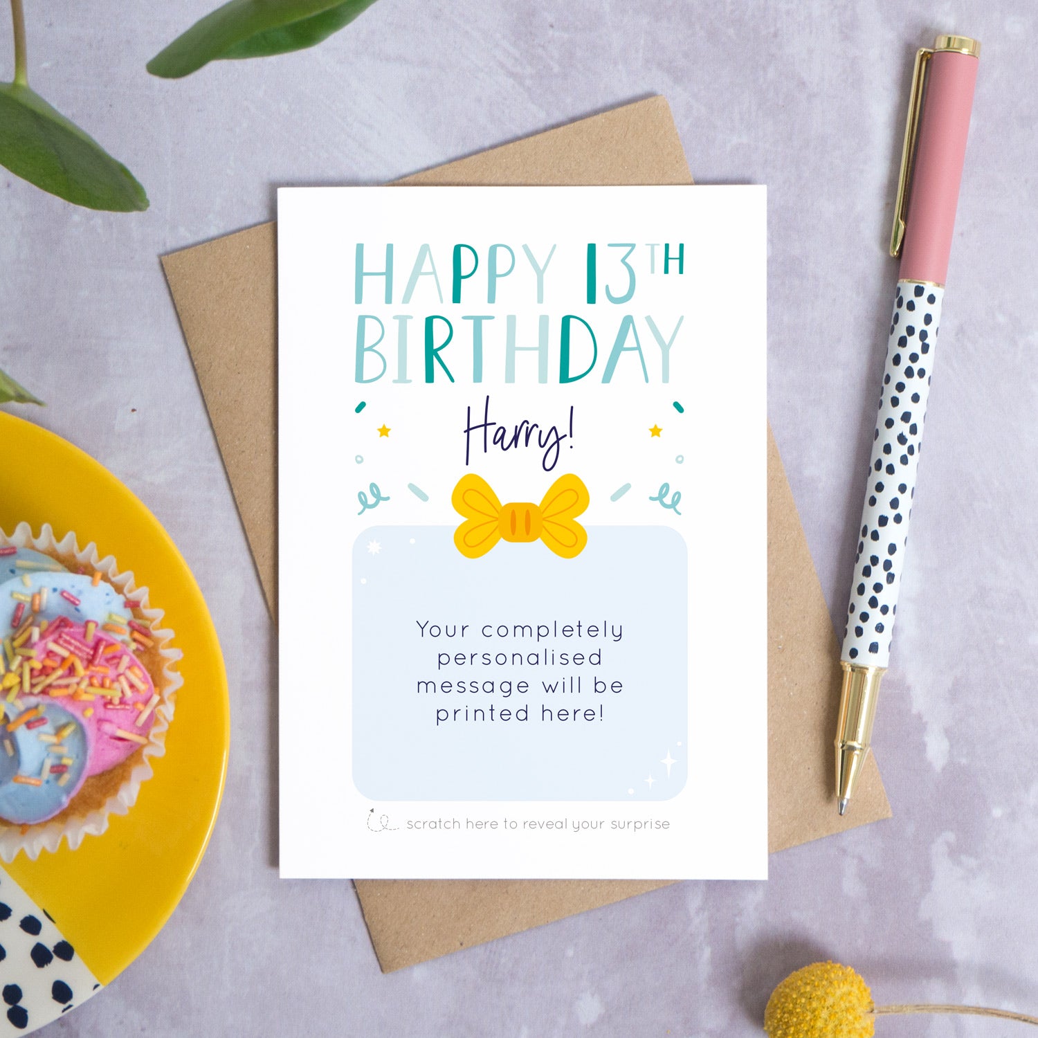 A personalised happy 13th birthday scratch card that has been photographed flat lay style on a grey concrete style background surrounded with foliage, a cupcake and a pen. The card itself shows how it would look once the gold panel has been completely removed. 