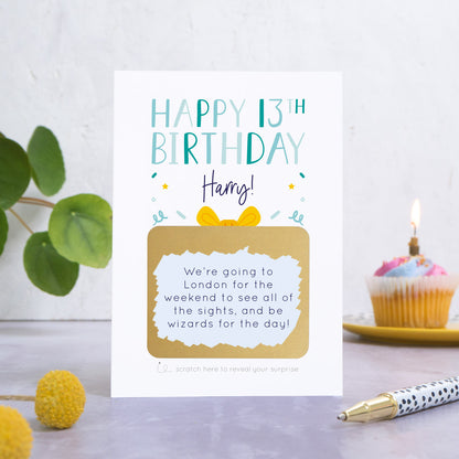 A personalised happy 13th birthday scratch card in blue that has been photographed on a white and grey background. There is foliage and yellow flowers on the left and a cupcake and a pen to the right. The card features the recipients name and a scratch panel that has been scratched off revealing a personalised message.