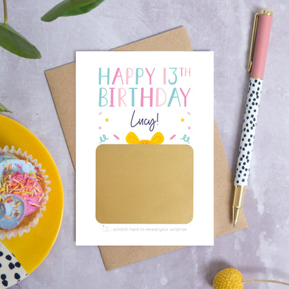 A personalised happy 13th birthday scratch card that has been photographed flat lay style on a grey concrete style background surrounded with foliage, a cupcake and a pen. The card itself shows how it will arrive and looks when it is completely unscratched.
