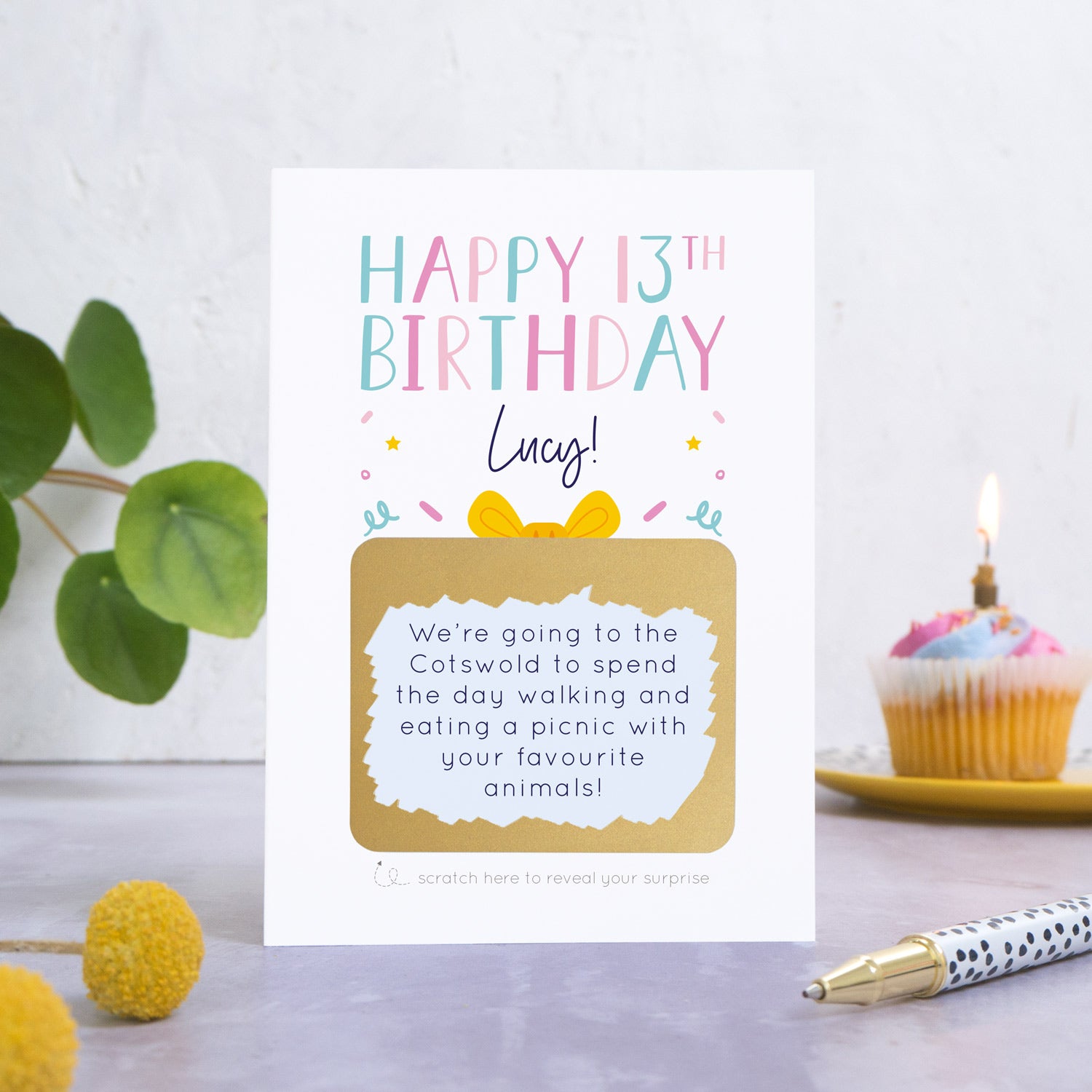 A personalised happy 13th birthday scratch card in pink that has been photographed on a white and grey background. There is foliage and yellow flowers on the left and a cupcake and a pen to the right. The card features the recipients name and a scratch panel that has been scratched off revealing a personalised message.