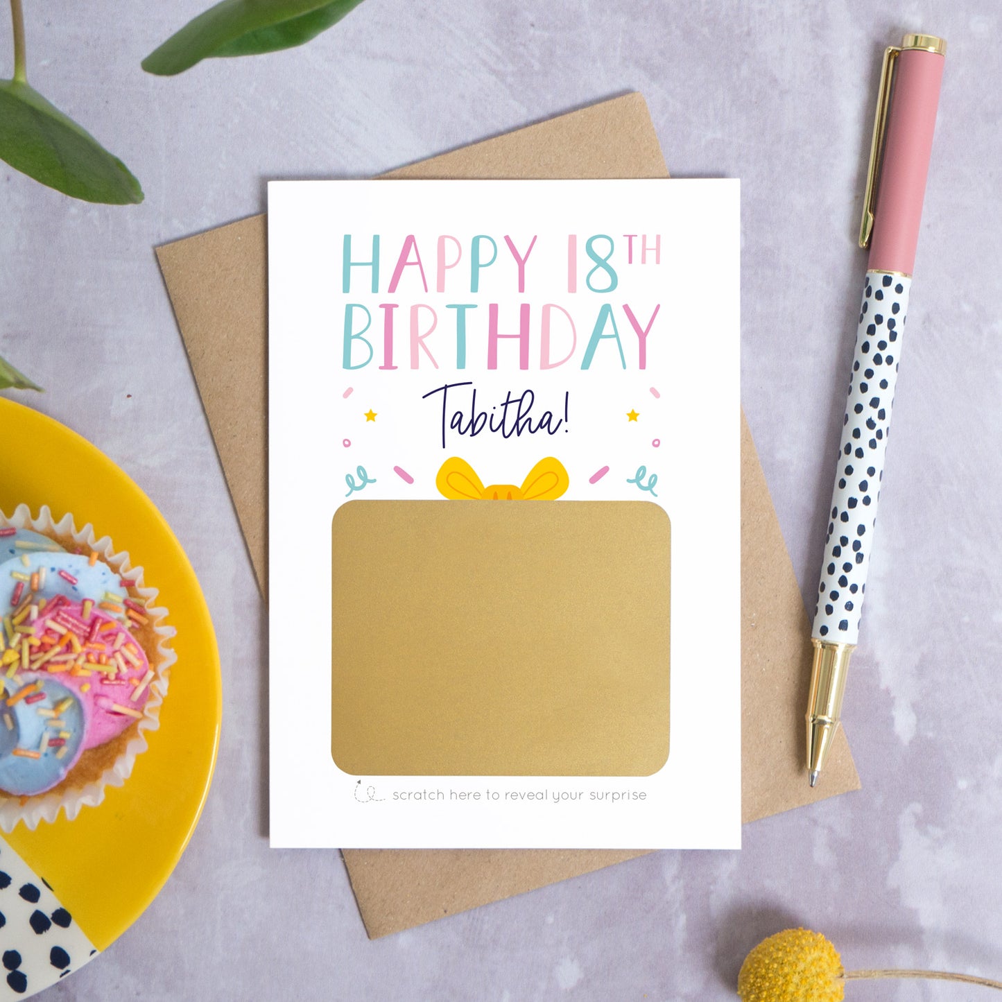 A personalised happy 18th birthday scratch card that has been photographed flat lay style on a grey concrete style background surrounded with foliage, a cupcake and a pen. The card itself shows how it will arrive and looks when it is completely unscratched.