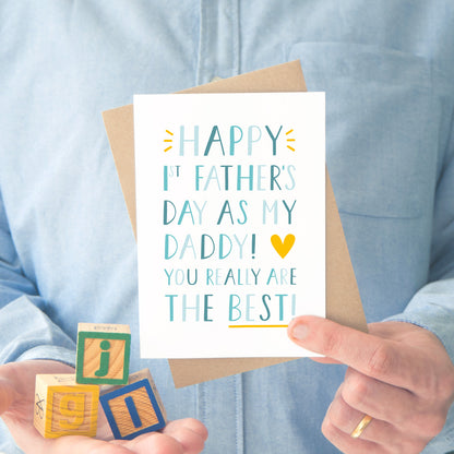 Happy 1st Father's Day as my daddy! You really are the best! A first father's day card photographed in front of a blue button up shirt with building blocks in the left hand and a the card and envelope being held in the right.