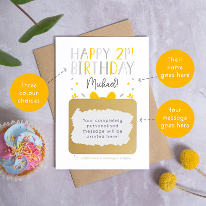 A personalised happy 21st birthday scratch card in grey that has been photographed on a grey background with foliage and a cupcake. There are also orange circles laid over the top of this image with arrows pointing to the areas that can be customised. In this instance they point to the name, the colour choices and the personalised scratch off message.