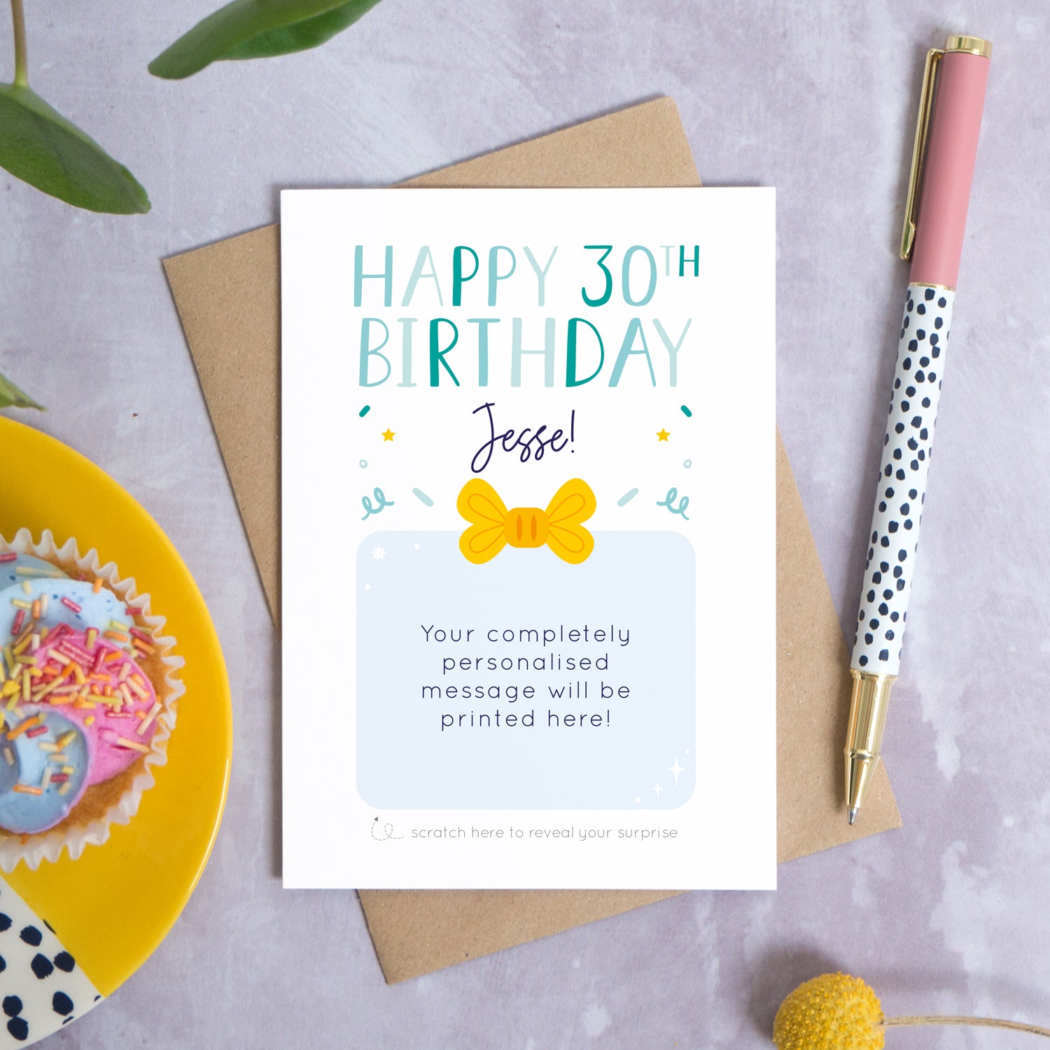 A personalised happy 30th birthday scratch card that has been photographed flat lay style on a grey concrete style background surrounded with foliage, a cupcake and a pen. The card itself shows how it would look once the gold panel has been completely removed. 