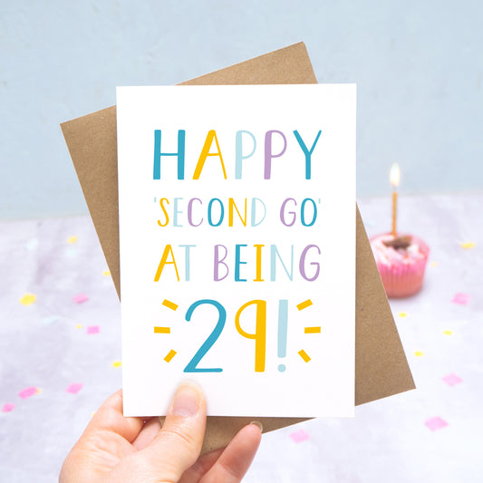 Happy second go at being 29 - milestone age card in blue, yellow and purple photographed on a grey and blue background with a cupcake and burning candle.