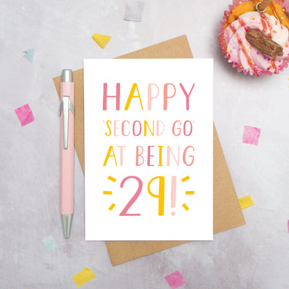 Happy second go at being 29 - milestone age card in pink photographed on a grey background surrounded by a cupcake, pen and confetti.