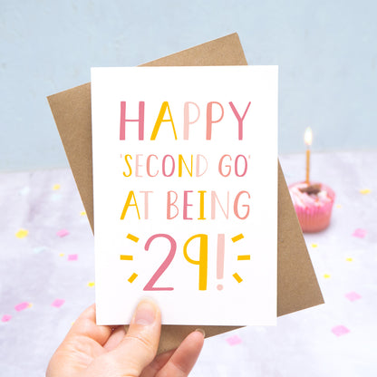 Happy second go at being 29 - milestone age card in pink photographed on a grey and blue background with a cupcake and burning candle.