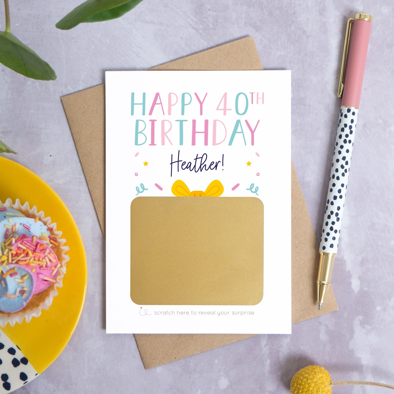 A personalised happy 40th birthday scratch card that has been photographed flat lay style on a grey concrete style background surrounded with foliage, a cupcake and a pen. The card itself shows how it will arrive and looks when it is completely unscratched.