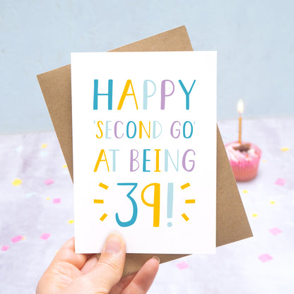Happy second go at being 39 - milestone age card in blue, yellow and purple photographed on a grey and blue background with a cupcake and burning candle.