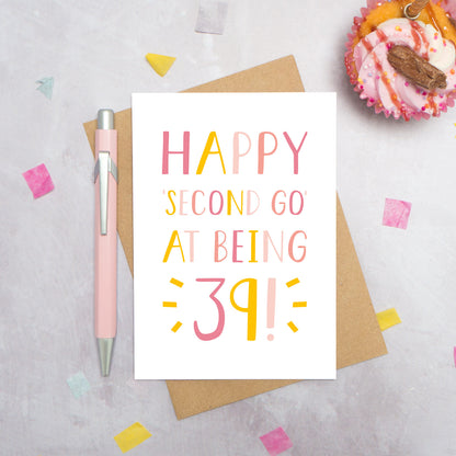 Happy second go at being 39 - milestone age card in pink photographed on a grey background surrounded by a cupcake, pen and confetti.