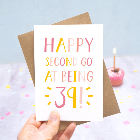 Happy second go at being 39 - milestone age card in pink photographed on a grey and blue background with a cupcake and burning candle.