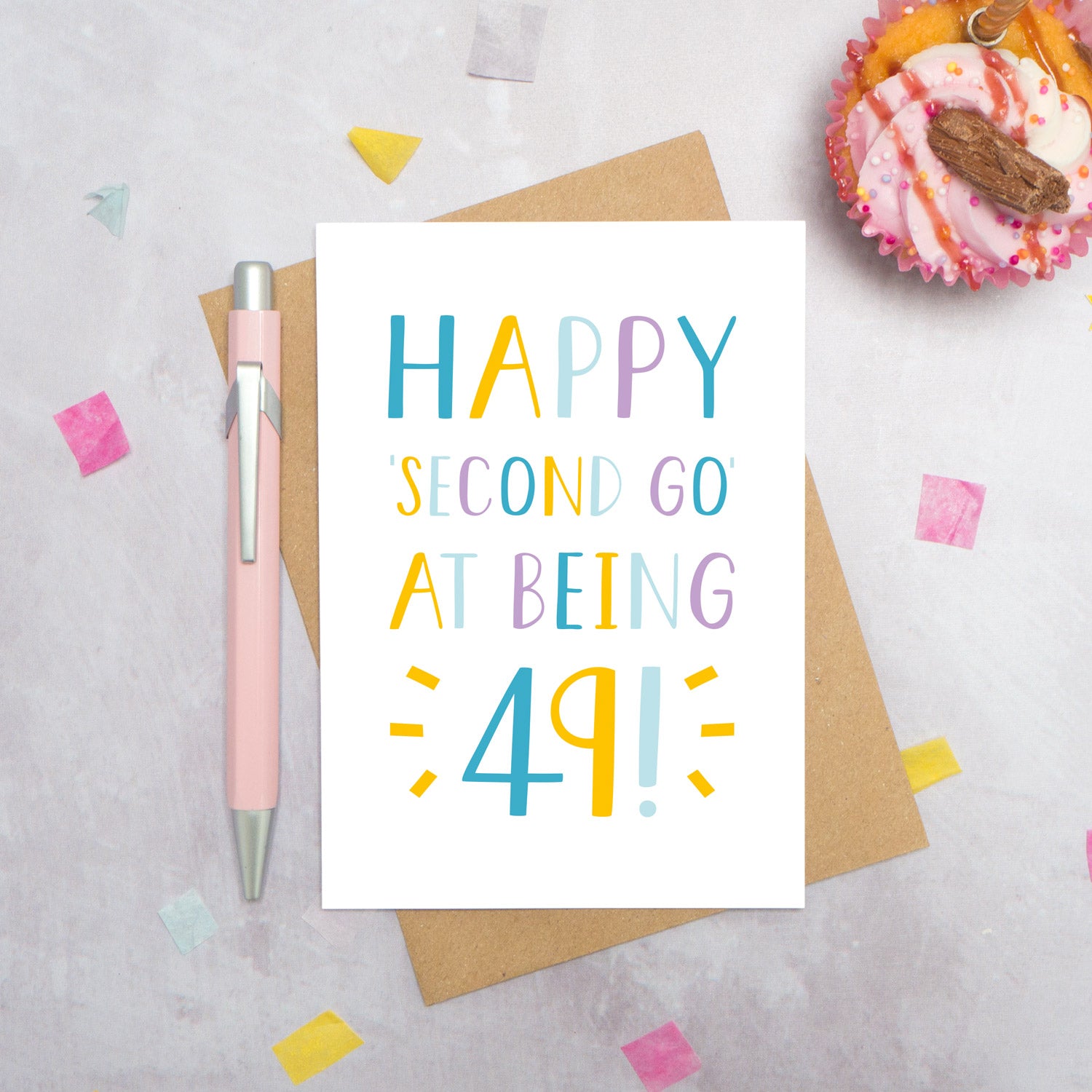 Happy second go at being 49 - milestone age card in blue, yellow and purple photographed on a grey background surrounded by a cupcake, pen and confetti.
